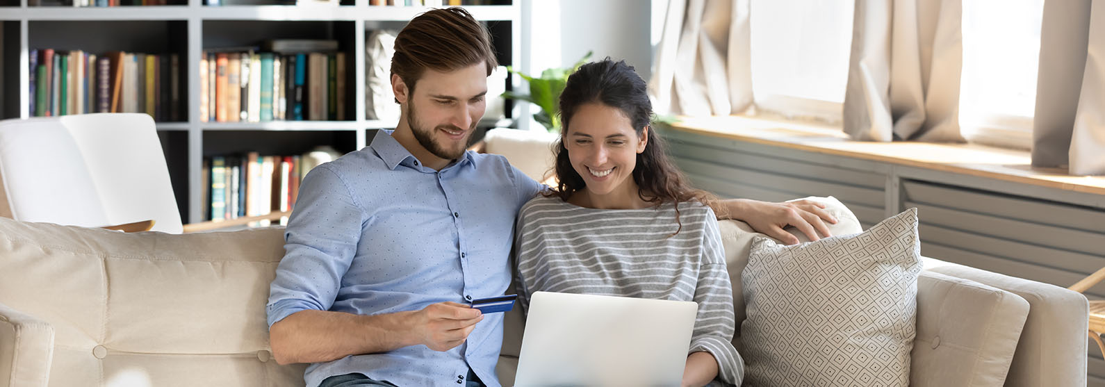 A man and woman sitting on a couch, holding a credit card, and looking at a laptop computer.