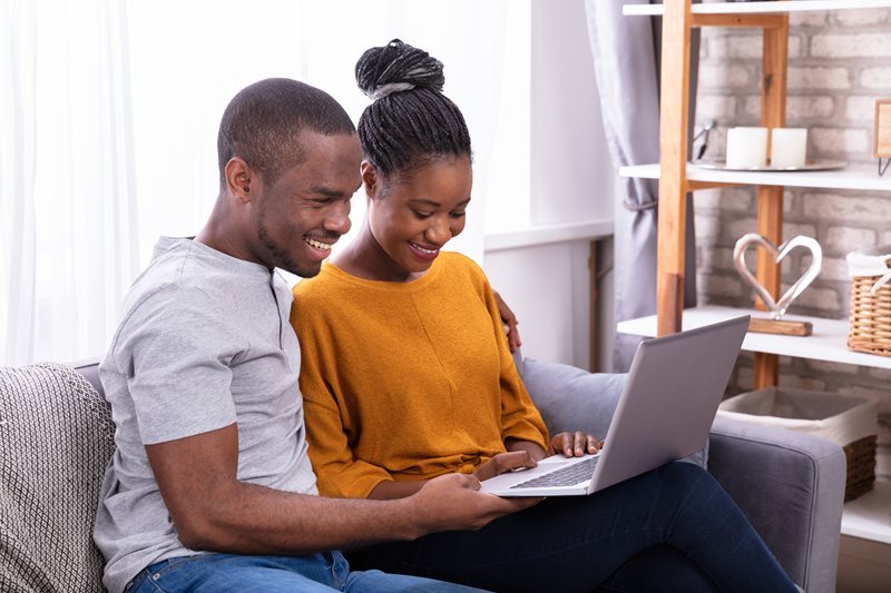 A man and woman, sitting on a couch, looking at their laptop computer.